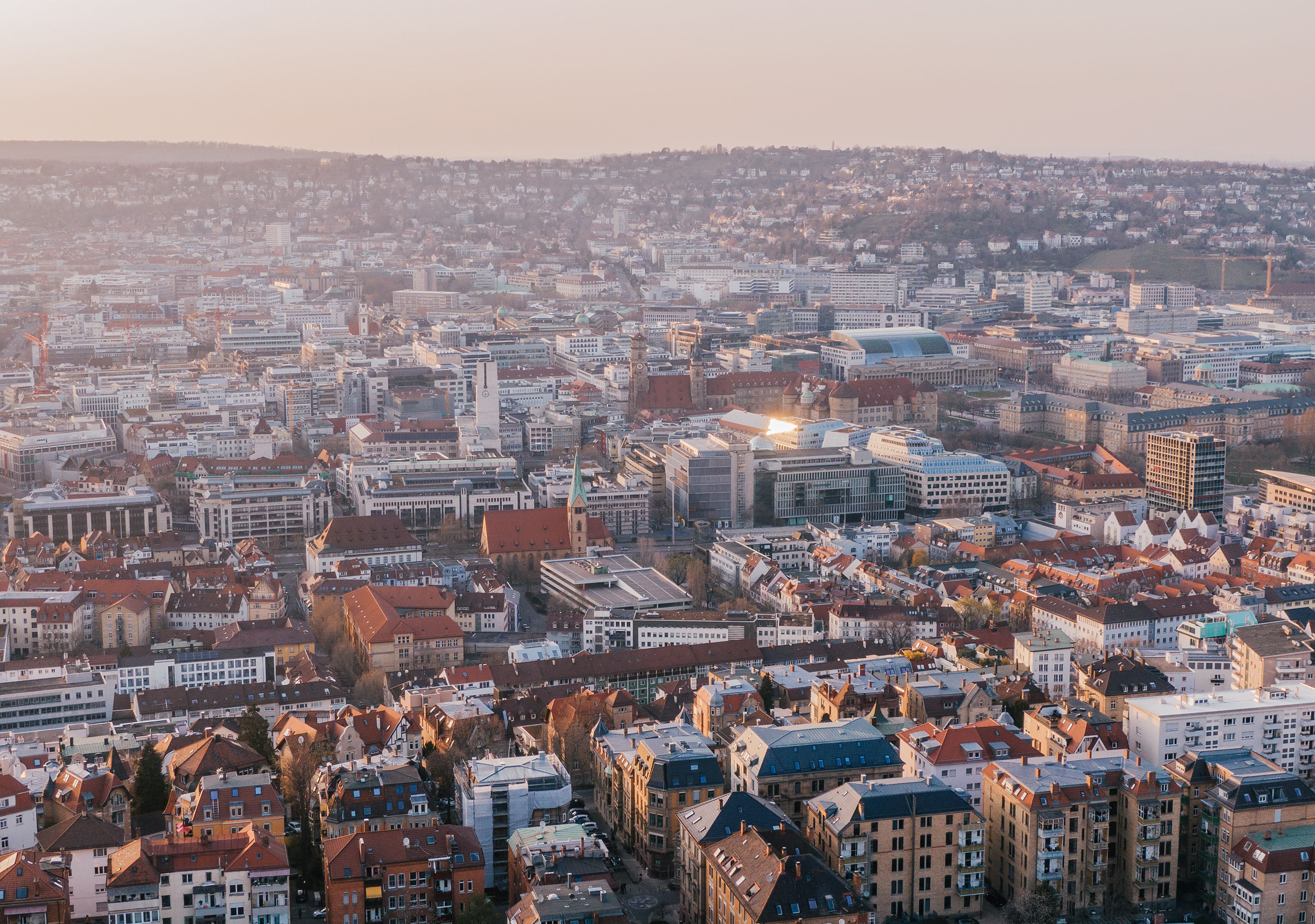 Luftaufnahme von Stuttgart bei Sonnenuntergang mit dicht bebauten Stadtvierteln, modernen und historischen Gebäuden. Im Hintergrund erstrecken sich Hügel mit Wohnhäusern.
