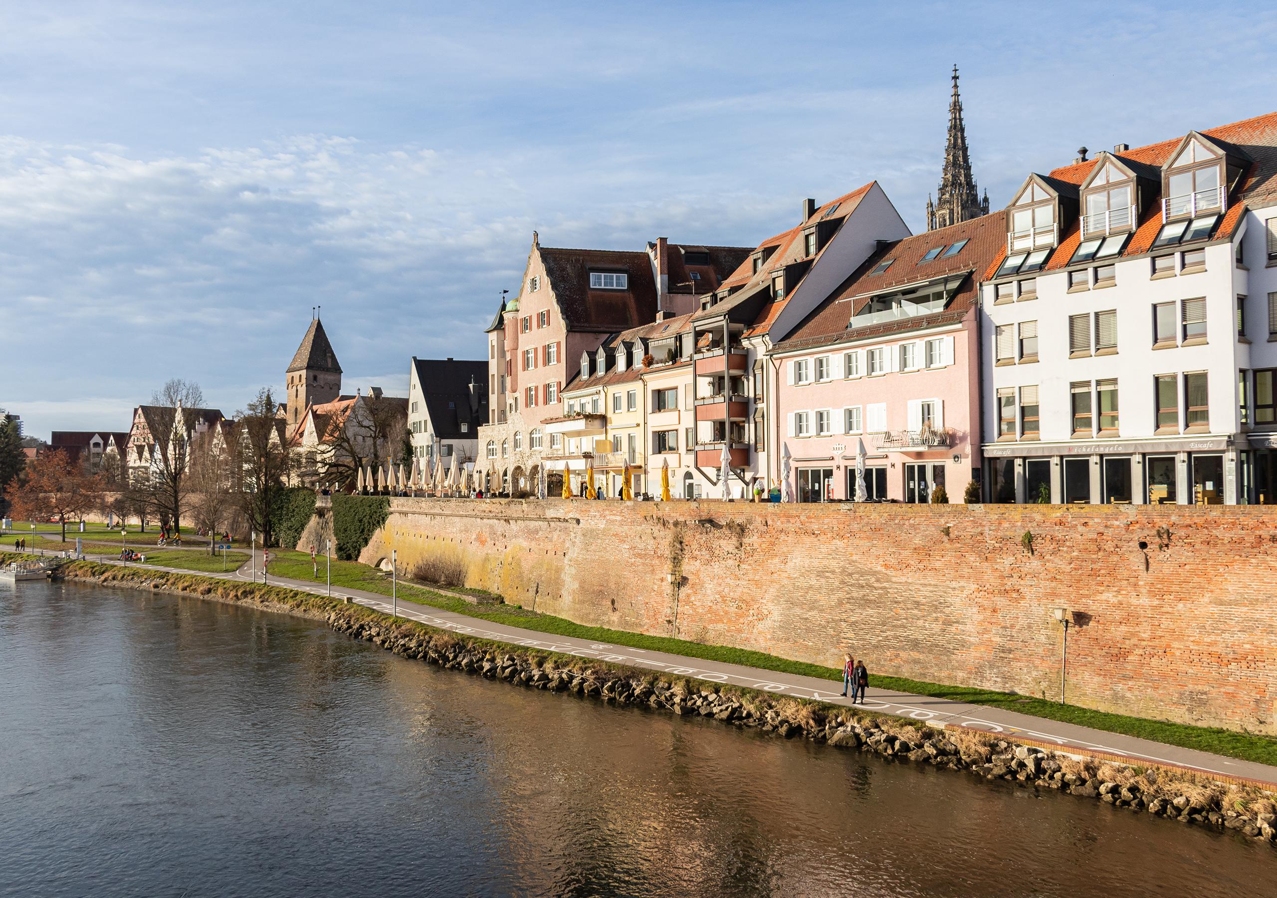 Blick auf die Altstadt von Ulm an der Donau mit historischer Stadtmauer, bunten Häusern und dem Ulmer Münster im Hintergrund. Fußgängerweg am Flussufer, beleuchtet von warmem Sonnenlicht.